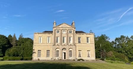 A large, classical-style house with ornate architectural details, surrounded by well-maintained greenery and a clear blue sky.